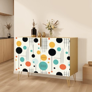 May include: A rectangular sideboard cabinet with gold-toned metal hairpin legs. The front doors feature a cream background with a repeating pattern of black, yellow, teal, and orange circles, thin vertical lines, and grid-like crosshatch designs.