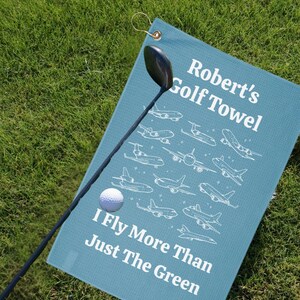Puede incluir: Una toalla de golf turquesa con el texto "Robert's Golf Towel" y "I Fly More Than Just The Green" en blanco. Presenta ilustraciones de aviones blancos. También se ven un palo y una bola de golf.