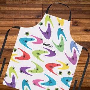 Könnte beinhalten: Weißer Küchenschürze mit einem bunten Retro-Bumerang-Muster. Die Schürze hat schwarze Träger und den Aufdruck "Heather". Geeignet zum Kochen oder Basteln, mit einem fröhlichen und farbenfrohen Muster.