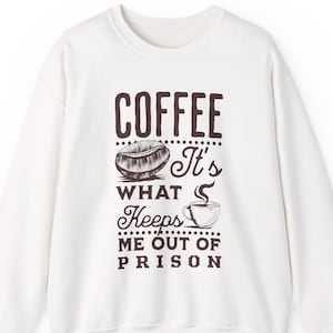 Peut inclure: Sweat-shirt blanc avec un motif sur le thème du café. Le design comprend le mot "COFFEE" au-dessus d'une illustration de grain de café, la phrase "It's What Keeps Me Out Of Prison" et une tasse de café.