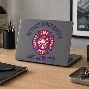 May include: A grey laptop displaying a sticker with a red and white firefighter emblem and the text "RETIRED FIREFIGHTER" and "CAPT. JOE WARREN" in blue. The laptop is open on a wooden desk.