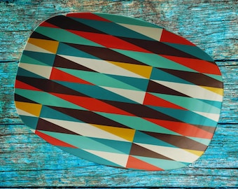 May include: An oval serving platter featuring a repeating geometric pattern of triangles in shades of teal, red, dark brown, yellow, and off-white. The platter has a smooth, flat surface and rounded edges, displaying a vibrant, multi-colored design.