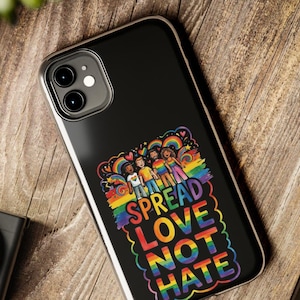 May include: Black phone case with a colorful graphic featuring a group of people, rainbows, and the words "SPREAD LOVE NOT HATE." The case has a silver border and is on a wooden surface.