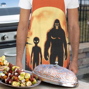 Puede incluir: Un delantal naranja con un diseño de Bigfoot y alienígena contra un fondo de puesta de sol. Una fuente plateada con tapa decorativa sobre una mesa, junto a una fuente de brochetas a la parrilla.