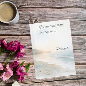 May include: A hardcover book with the title "Writings from the heart..." and the name "Hannah" on the cover. The cover features a watercolor beach scene. A cup of coffee and pink flowers are next to the book on a wooden surface.