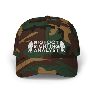 May include: A camouflage baseball cap with the text "BIGFOOT SIGHTING ANALYST" embroidered in white on the front.