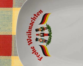 May include: White oval plate with the words "Frohe Weihnachten" in red script. The plate features three toy soldier figures, German flags, and a green wreath design. The plate is on a red and yellow checkered surface.