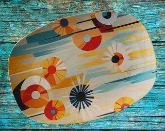May include: An oval serving platter featuring a graphic pattern of stylized circular sunbursts and floral shapes in shades of orange, red, yellow, teal, and black, set against a light cream background with horizontal streaks of color.
