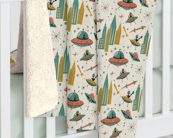 May include: A cream-colored blanket with a retro space-themed pattern. The design features flying saucers, aliens, skyscrapers, and starbursts in teal, orange, and gold. The blanket has a soft, cream-colored sherpa lining.