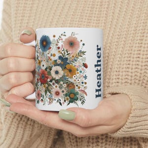 Puede incluir: Taza de cerámica blanca con un diseño floral colorido con flores azules, rosas, amarillas y rojas. La palabra "Heather" está impresa verticalmente en azul. La taza es sostenida por una persona.