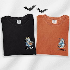 May include: A black t-shirt and an orange t-shirt, each featuring a graphic of a ghost wearing a hat and holding a pumpkin bucket amidst spiderwebs and bats. The black shirt has the name OLIVIA printed below, and the orange shirt has DANIEL.