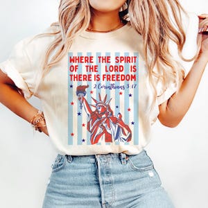 May include: Cream-colored t-shirt featuring a graphic of the Statue of Liberty in red, white, and blue, holding a torch. The text reads "WHERE THE SPIRIT OF THE LORD IS THERE IS FREEDOM 2 Corinthians 3:17" with stars and stripes.