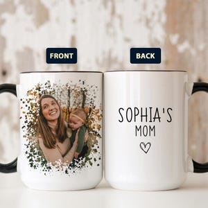 May include: White ceramic mug with a black handle and rim. The front features a photo of a woman and baby surrounded by a watercolor-style design. The back of the mug reads "Sophia's Mom" with a heart symbol.