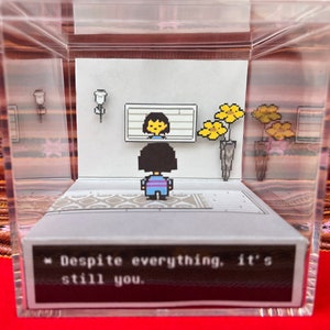 May include: A clear acrylic box with a diorama inside depicting a video game character standing in front of a mirror. The character is a young person with brown hair and a blue and purple striped shirt. The text "Despite everything, it's still you." is displayed on a black background at the bottom of the diorama.