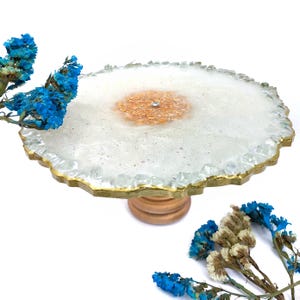 Pode incluir: Um suporte decorativo para bolos com um tampo de resina branco e dourado. O tampo tem um centro de glitter dourado e uma borda pintada de ouro. O suporte é suportado por uma base de madeira. Flores azuis secas em primeiro plano.
