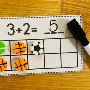May include: A dry erase board with a math problem, 3 + 2 = 5, and a grid with sports-themed counters. There are three basketballs, one soccer ball, and one tennis ball. A black dry erase marker is next to the board.