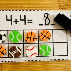 May include: A white dry-erase board with a math problem written on it: 4 + 4 = 8. There are eight colorful sports-themed erasers on the board, including soccer balls, basketballs, baseballs, and tennis balls.