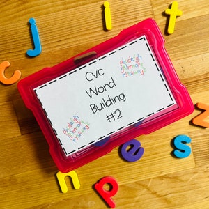 May include: A pink plastic container with a white label that says "Cvc Word Building #2". The container is filled with colorful foam letters. There are also foam letters scattered around the container on a wooden surface.