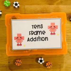 May include: An orange plastic container with a white label that says "Tens Frame Addition". The label has a cartoon plus sign with a smiling face. There are several colorful erasers around the container, including a football, a tennis ball, and basketballs.