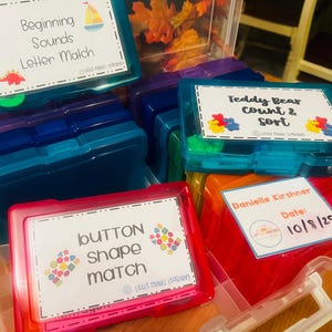 Op de afbeelding: Een verzameling kleurrijke plastic bakjes met labels voor educatieve activiteiten. De labels luiden "Beginning Sounds Letter Match", "Teddy Bear Count & Sort" en "Button Shape Match". De bakjes zijn in verschillende kleuren, waaronder blauw, rood en roze.