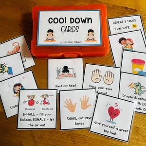 Może przedstawiać: Pomarańczowe plastikowe pudełko z "COOL DOWN CARDS" i różnymi ilustrowanymi kartami. Karty zawierają instrukcje, takie jak "Rest my head", "Close your eyes" i "Give yourself a great big hug!" dla uspokajających aktywności. Karty leżą na drewnianej powierzchni.