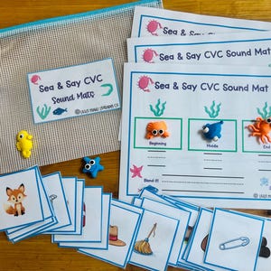 May include: A set of Sea & Say CVC Sound Mats with blue borders, featuring illustrations of sea creatures. Includes small clay figures of a crab, fish, and octopus, a mesh storage pouch, and a stack of square cards depicting a fox, broom, and pin.