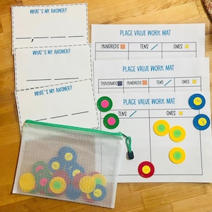 May include: Educational math set featuring place value work mats, question cards, and colorful counters. The mats display place value columns for ones, tens, hundreds, and thousands. A clear mesh bag contains various colored counters for hands-on learning.