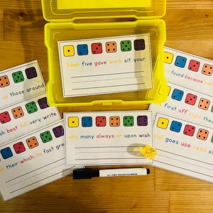 May include: A yellow plastic box contains flashcards featuring dice graphics and sight words. Several cards are arranged around the box, alongside a dry-erase marker and a small yellow die. The cards are designed for educational purposes.