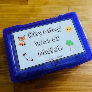 以下が含まれることがあります： 「Rhyming Words Match」というタイトルの学習ゲームが入った青いプラスチック製の容器。ゲームの表紙には、漫画のキツネ、靴下、太陽、木が描かれています。このゲームは教育目的で設計されています。