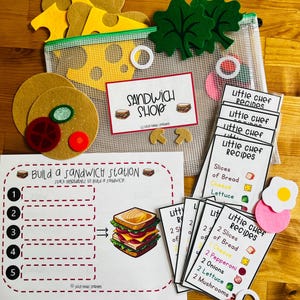 Könnte beinhalten: Ein Kinderspiel-Lebensmittelset mit Filz-Sandwich-Zutaten, darunter Brot, Käse, Salat und Tomatenscheiben. Das Set enthält Rezeptkarten und ein "Sandwich Shop"-Schild, das fantasievolles Spielen und Lernen fördert.
