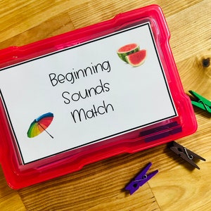 May include: A pink plastic storage box with a white label that says "Beginning Sounds Match". The label also has a picture of a rainbow umbrella and a slice of watermelon. There are three colorful clothespins on a wooden surface.