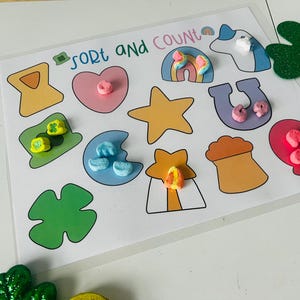May include: A learning activity with the words "Sort and Count" printed on a white background. The activity features colorful shapes with matching marshmallow pieces, including a rainbow, horseshoe, and star. Green glitter shamrocks are also visible.