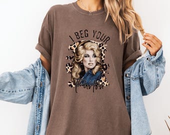 May include: A brown t-shirt with a graphic of Dolly Parton in a leopard print frame. The text "I Beg Your Pardon" is printed on the shirt.