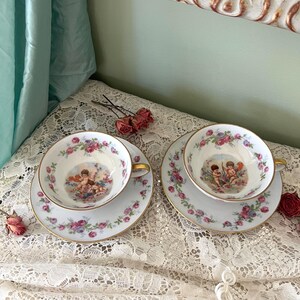Puede incluir: Dos tazas de té y platillos de porcelana blanca con ribetes dorados. Las tazas tienen un patrón floral y una imagen central de dos querubines. Los platillos tienen un patrón floral con flores rosas, moradas y azules.
