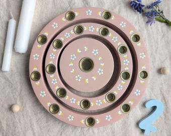 May include: A pink wooden menorah with three concentric circles, each with small candle holders. The circles are decorated with white and yellow flowers.