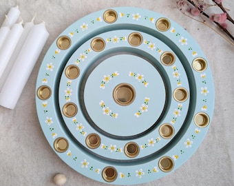 May include: A pale blue wooden candle holder with three circular tiers, each with gold candle holders. The holder is decorated with small white and yellow floral patterns. White candles are visible to the left.