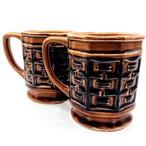 May include: Two brown ceramic mugs with a geometric pattern. The mugs have a dark brown design on a lighter brown background. Each mug has a handle and a slightly flared rim. The mugs are likely for coffee or tea.