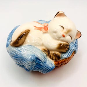 Puede incluir: Una figura de cerámica de un gato siamés durmiendo con marcas marrones, un lazo rosa y los ojos cerrados. El gato está acurrucado sobre un cojín azul dentro de una cesta tejida. La figura está pintada con colores brillantes y brillantes.