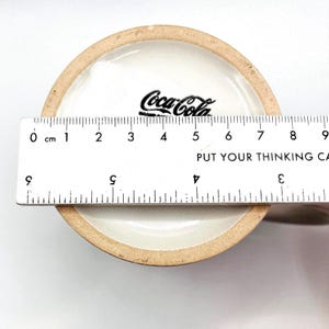 May include: A ceramic Coca-Cola mug, viewed from the inside bottom. The Coca-Cola logo is printed in black. A ruler is placed across the mug's opening, showing a measurement of approximately 9 cm. The text "PUT YOUR THINKING CA" is visible.