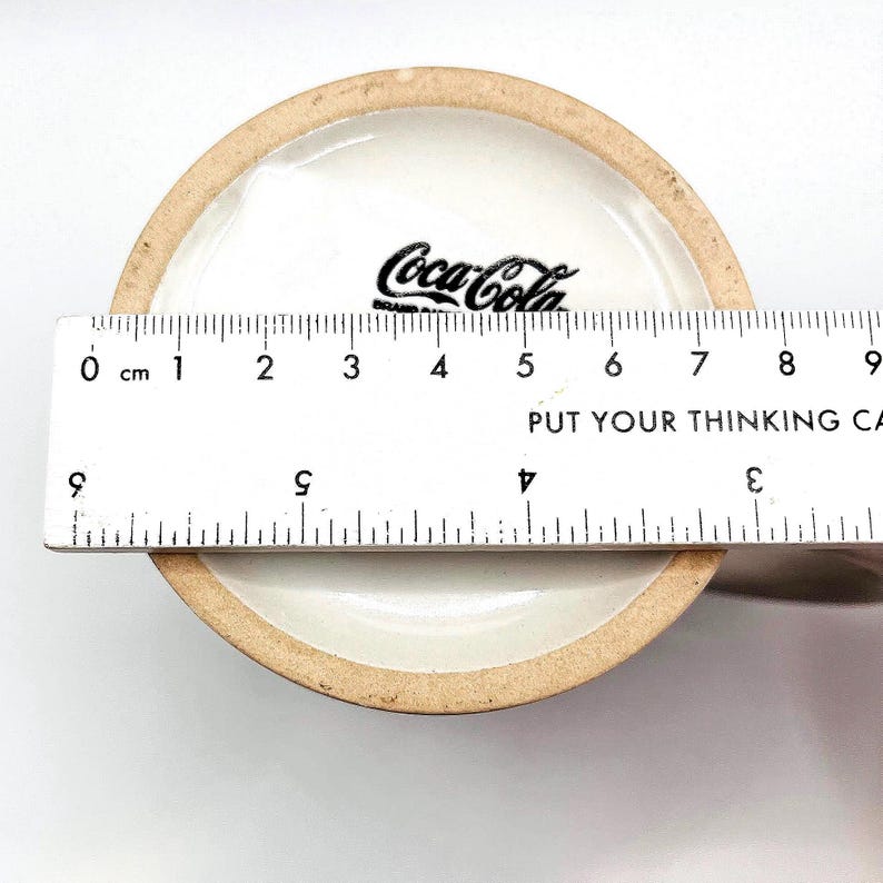 May include: A ceramic Coca-Cola mug, viewed from the inside bottom. The Coca-Cola logo is printed in black. A ruler is placed across the mug's opening, showing a measurement of approximately 9 cm. The text "PUT YOUR THINKING CA" is visible.