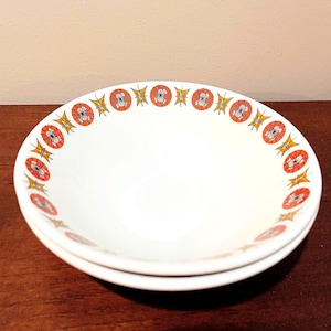 Peut inclure: Deux bols en céramique blanche empilés. Chaque bol présente une bordure décorative de motifs floraux orange et rouges avec des accents jaunes. Les bols sont sur une surface en bois foncé.