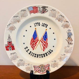 Può includere: Un piatto commemorativo del bicentenario americano. Il piatto in ceramica bianca presenta gli anni 1776-1976, due bandiere americane incrociate e un calendario sul bordo. Il piatto include anche illustrazioni di personaggi e monumenti storici.