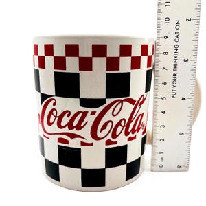 May include: A white ceramic Coca-Cola mug with a black and red checkerboard pattern. The Coca-Cola logo is in red. The mug is approximately 4 inches tall.