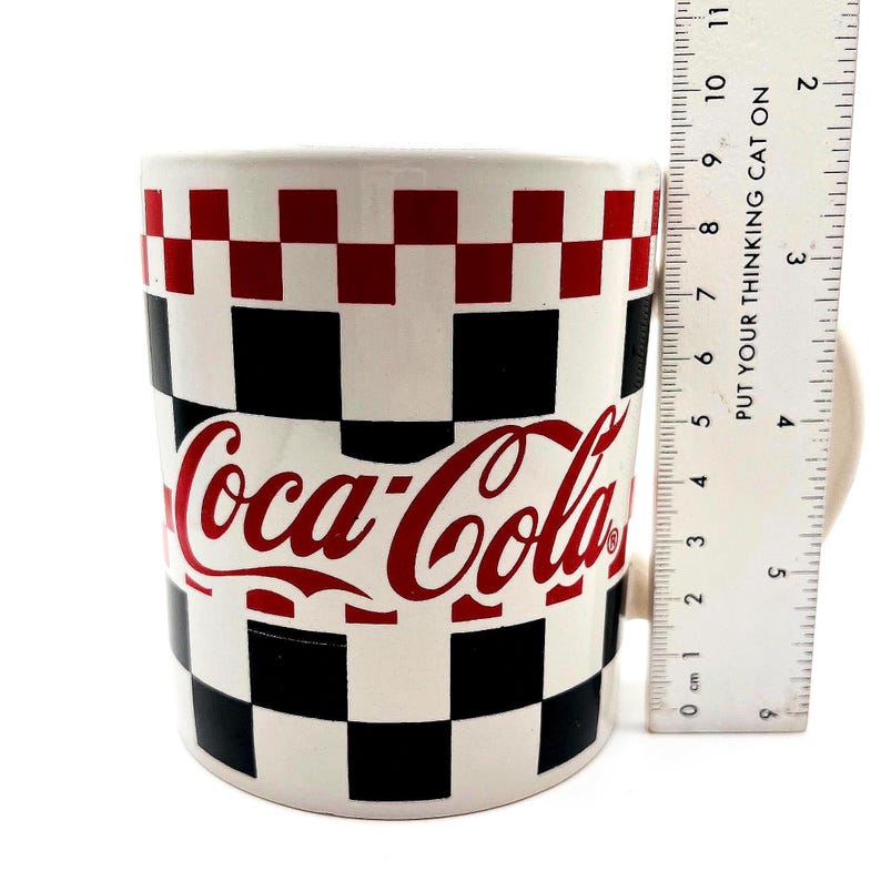 May include: A white ceramic Coca-Cola mug with a black and red checkerboard pattern. The Coca-Cola logo is in red. The mug is approximately 4 inches tall.