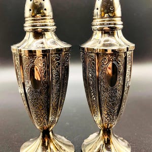 Puede incluir: Un par de saleros y pimenteros plateados. Cada salero tiene un diseño ornamental con un patrón floral. Los saleros tienen un cuerpo bulboso, una base acampanada y una parte superior perforada. El fondo es oscuro.