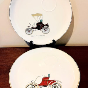 Puede incluir: Dos platos decorativos blancos con bordes negros y ribete dorado. Cada plato presenta una ilustración de coche antiguo. Uno muestra un coche negro con techo beige, etiquetado "Winton Automobile, 1898". El otro muestra un coche rojo, etiquetado "Oldsmobile Runabout, 1903."