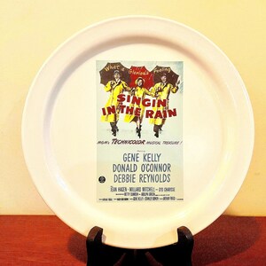 May include: A white ceramic plate featuring a vintage movie poster for "Singin' in the Rain." The poster shows Gene Kelly, Donald O'Connor, and Debbie Reynolds. The plate is on a black stand.