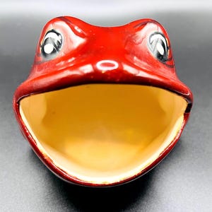 May include: A ceramic frog-shaped bowl with a wide open mouth. The bowl is painted a glossy red on the exterior and has a yellow interior. The frog's eyes are white with black pupils. The bowl is designed to resemble a frog's head.