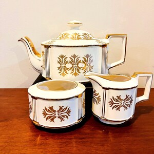 May include: A white tea set with gold detailing, comprising a teapot, creamer jug, and sugar bowl. The set features a gold floral pattern and gold trim. The teapot has a lid and handle. The creamer has a spout and handle. The sugar bowl has a lid.
