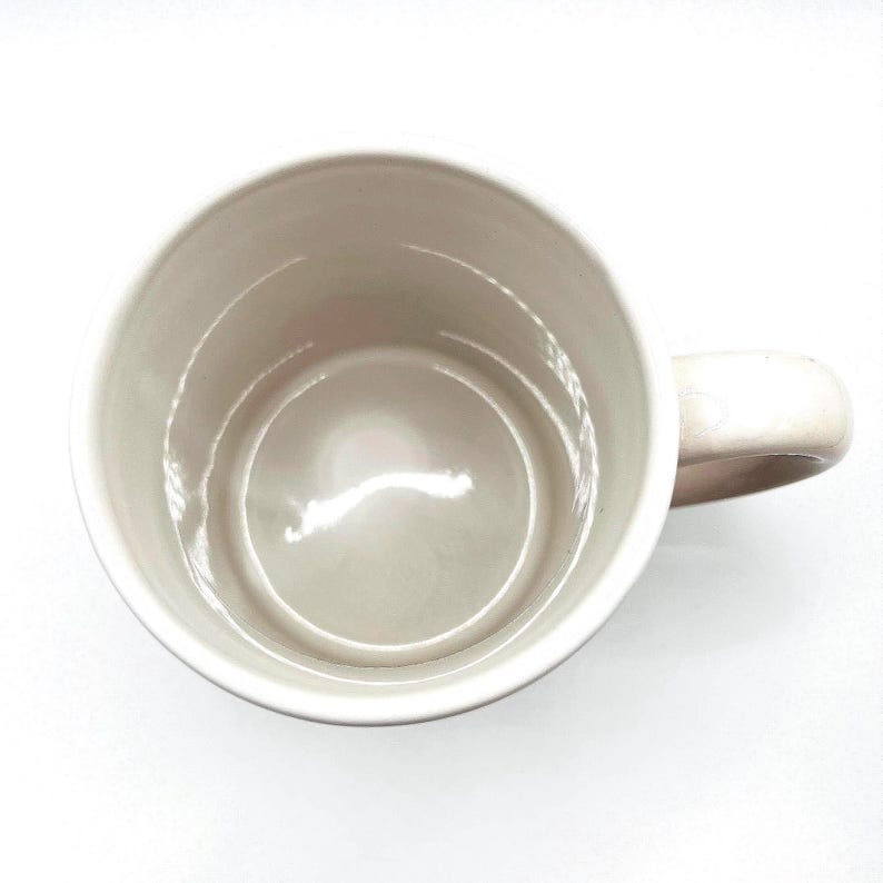 May include: A beige ceramic mug with a rounded handle, viewed from above. The interior of the mug is a lighter shade of beige, and the handle is on the right side of the image. The mug is set against a white background.